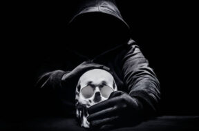 Horror photo of a scary man in hoodie sitting and silencing human skull.