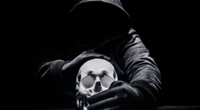 Horror photo of a scary man in hoodie sitting and silencing human skull.